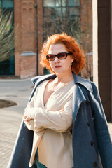 Attractive red-haired woman in an oversized shirt and coat thrown over her shoulders stands against the backdrop of business district. Vertical portrait of woman with her arms folded across her chest