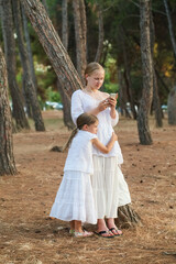 Two sisters, kid and teenage girl, younger one hugging older one which is holding smartphone, they standing near tree in the park