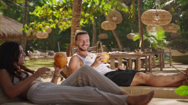 Young happy multiethnic couple relaxing on sunbeds at tropical resort clink and drink coconut water. Cheerful biracial tourists on summer vacation having fresh king coconuts with exotic background