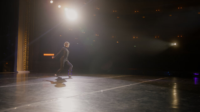 Female dancer performs on theater stage illuminated by spotlights. Ballerina in training suit dances and practices graceful ballet movements at choreography rehearsal. Art of modern ballet dance.