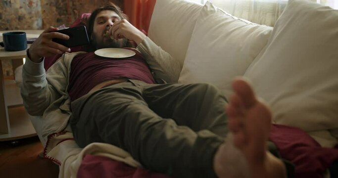 Carefree, careless lazy beard man eating sandwich, watching some entertaining content on his smartphone, laughing, lying on the couch. Eating disorder, overweight. Laziness, procrastination concept.