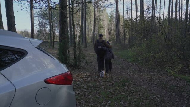 Father and son leave parked car and walk hugging along path from auto into forest. Little dog in nice overalls runs after owners in autumn forest