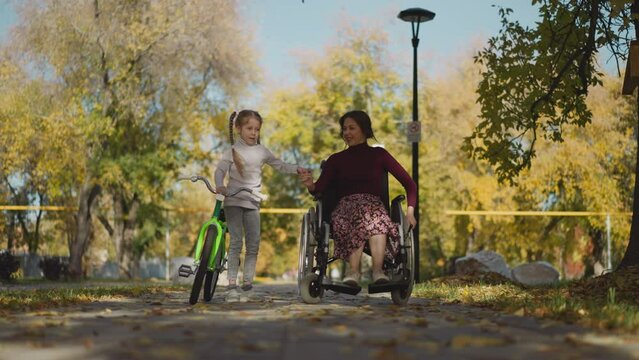 Mother with disability in wheelchair and preschooler daughter with bicycle rest together in park. Smiling couple moves in relaxed manner holding hands in caringly