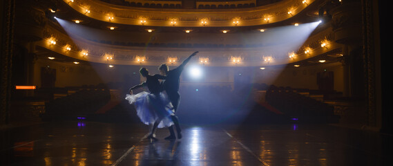 Couple of classical ballet dancers practice on theatre stage before performance. Man in training suit lifts graceful ballerina. Dance choreography rehearsal. Illuminated theatrical hall. Slow motion. © Framestock