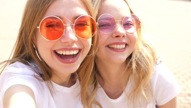 Two young smiling hipster blond women in summer white t-shirt clothes. Girls taking selfie self portrait photos on smartphone.Models posing on street background.Female showing positive emotions