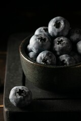 Vertical close-up view of a bowl of blueberries on the wooden board before the dark background