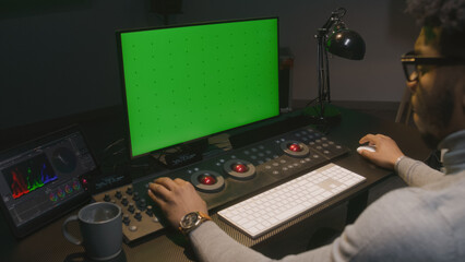 African American film maker works on computer in studio with digital equipment. Color grading control surface and tablet with professional software standing on the table. PC monitor with green screen.