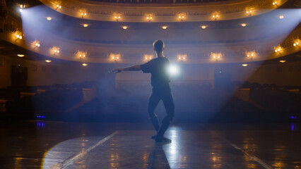 Ballet dancer in training suit practices visual moves on theater stage and prepares to performance. Dancer on choreography rehearsal before show. Illuminated theatrical hall. Classical ballet art.