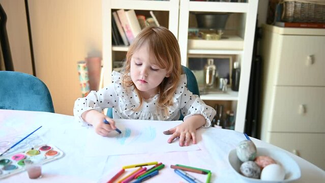 Small girl sitting at the diner table, making drawings and being creative. She is painting with a blue crayon on a piece of paper. 4K