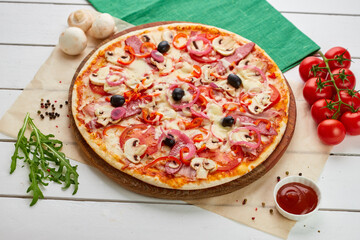 Freshly baked pizza with smoked sausages, red pepper, mushrooms, onion and olives served on wooden background with tomatoes, sauce and herbs. Food delivery concept. Restaurant menu