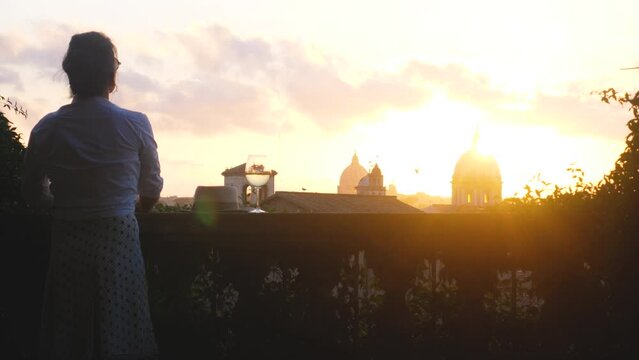 Young woman tourist fashion white dress with glass of white wine in front of panoramic view of Rome cityscape from campidoglio terrace at sunset. Landmarks, domes of Rome, Italy.