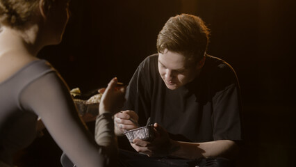 Pair of tired ballet dancers sit on theatre stage and discuss performance. Ballerina and dance partner eat together after choreography rehearsal. Empty theatrical hall. Classical ballet art concept.