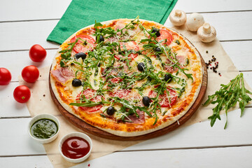 Freshly baked pizza with ham, rukkola, sauce pesto and olives served on wooden background with tomatoes, sauces and herbs. Food delivery concept. Restaurant menu