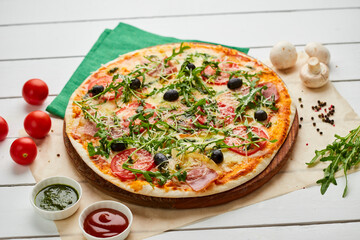 Freshly baked pizza with ham, rukkola, sauce pesto and olives served on wooden background with tomatoes, sauces and herbs. Food delivery concept. Restaurant menu