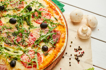 Freshly baked pizza with ham, rukkola, sauce pesto and olives served on wooden background with tomatoes, sauces and herbs. Food delivery concept. Restaurant menu