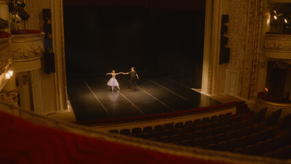 Establishing shot of ballet dancers preparing theatrical dance performance. Man and woman practice choreography on classic theater stage. Classical ballet dance. Rows of seats. Dramatic lighting.