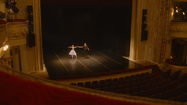 Establishing shot of ballet dancers preparing theatrical dance performance. Man and woman practice choreography on classic theater stage. Classical ballet dance. Rows of seats. Dramatic lighting.
