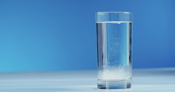 Close-up of a glass and fingers tossing a white round soluble aspirin tablet bubbling in a glass of water against a blue background. Acetylsalicylic acid effervescent tablet dissolves in water.