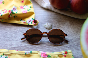 Floral yellow bikini, wicker bag, retro sunglasses, seashell, bag with peaches and cut watermelon on wooden background. Selective focus.