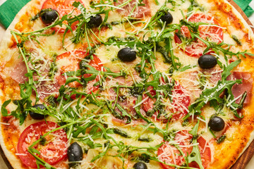 Freshly baked pizza with ham, rukkola, sauce pesto and olives served on wooden background with tomatoes, sauces and herbs. Food delivery concept. Restaurant menu