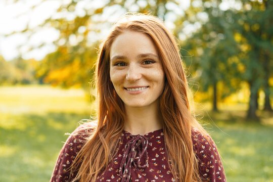 Outdoors portrait of a young beautiful red head smiling woman in sunlight in a green park.