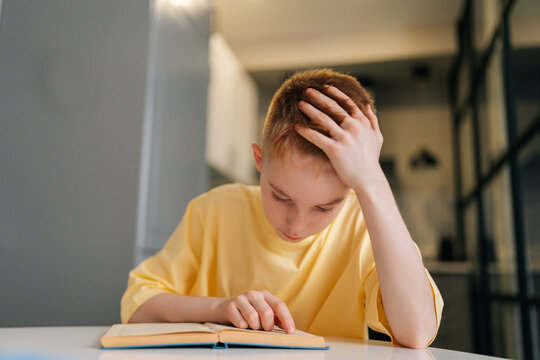 Closeup of tired pupil schoolboy reading paper book following lines with finger along page sitting at table in living room. Portrait of little student boy learning at home with textbook.