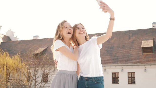Two young smiling hipster blond women in summer white t-shirt clothes. Girls taking selfie self portrait photos on smartphone. Models posing on street background. Female showing positive face emotions