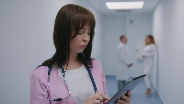 A nurse holds a tablet while studying a patient's medical record while standing in the hallway