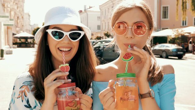 Two young beautiful smiling hipster girls in trendy summer clothes. Carefree women posing in the street. Positive models having fun in sunglasses. Drinking fresh cocktail drink