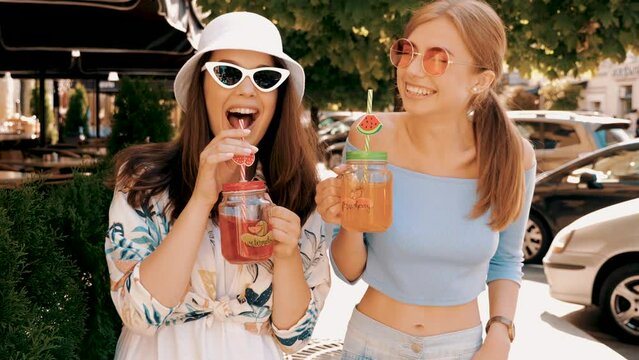 Two young beautiful smiling hipster girls in trendy summer clothes. Carefree women posing in the street. Positive models having fun in sunglasses. Drinking fresh cocktail drink