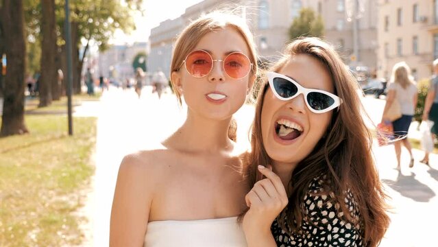 Portrait of two young beautiful smiling hipster girls in trendy summer clothes. Carefree women posing on street background. Positive models having fun in sunglasses and blowing bubble with chewing gum