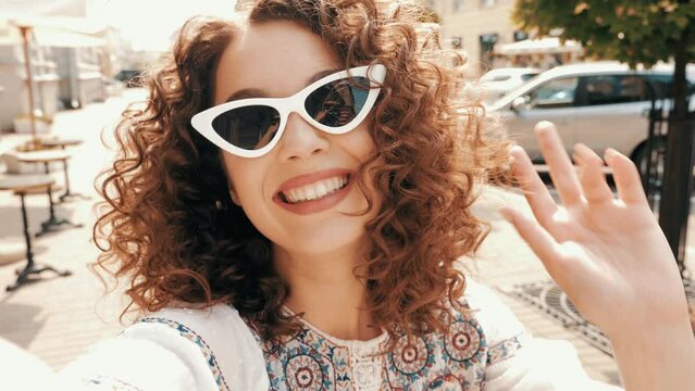 Beautiful smiling model with afro curls hairstyle dressed in summer hipster white dress. Sexy carefree woman posing in the street in sunglasses. Taking selfie self portrait photos on smartphone
