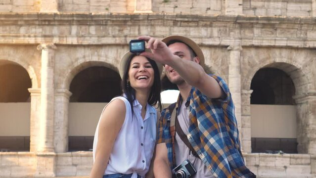 Lovely couple of tourist sitting on a wall at Colosseum taking selfie videos with action camera. Rome, Italy