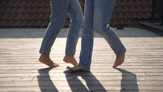 Feet of woman and man in jeans barefoot playfully and cheerfully dance on their toes, fool around and step on each other feet in sunlight. Terrace with wooden floor and shadows from couple in love.