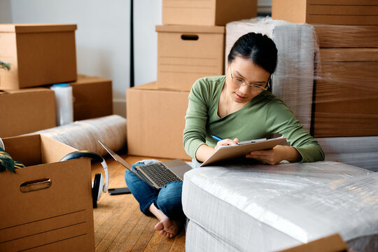 Asian Woman Going Through Checklist While Using Laptop And Moving Into New Home.