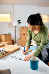 Asian woman writes list of her belongings while packing to move into new apartment.