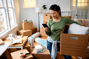 Happy Asian woman texting on cell phone while moving into new apartment.