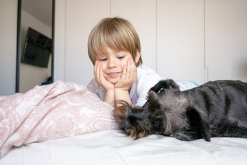 Cute blond child, toddler boy, lying on the bed with his pet dog at home