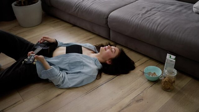 Young woman playing ukulele at home while laying on the floor near sofa