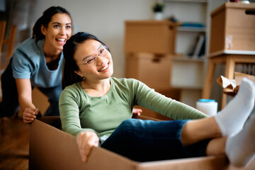 Happy Asian woman has fun while being pushed by her female friend in cardboard box after moving into new apartment.
