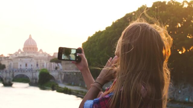 Young blonde woman tourist with hat on a bridge on river tiber in Rome, taking selfies with smartphone. View of of St. Peter and Vatican city, Italy.