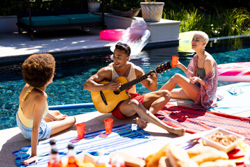 Happy diverse group of friends having pool party, playing guitar in garden