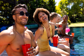 Happy diverse group of friends having pool party, holding plastic cups in garden