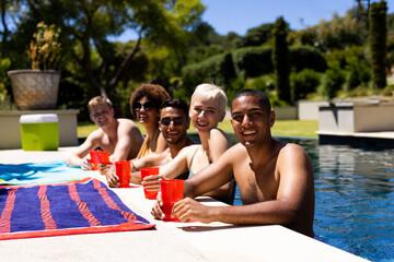 Portrait of happy diverse group of friends having pool party, holding plastic cups in garden
