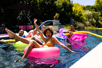 Happy diverse group of friends having pool party, using swim rings in swimming pool in garden