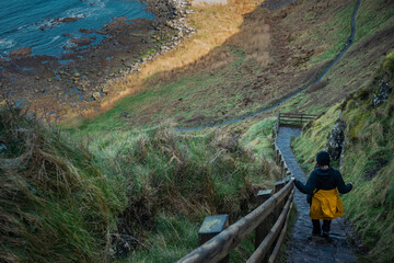 Woman descending towards hexagonal stones or pillars at Giants causeway in northern ireland, majestic basalt pillars at the beach on a cloudy day. Wide view of area with stairs.