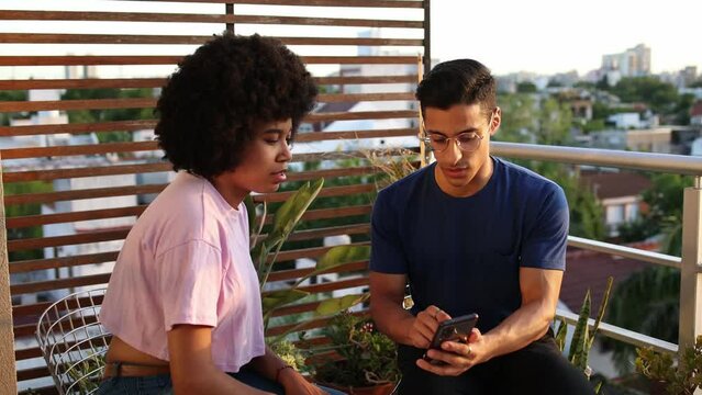 Attractive Hispanic man on a balcony wearing a blue shirt explaining content on his mobile phone to a friend with afro hairstyle