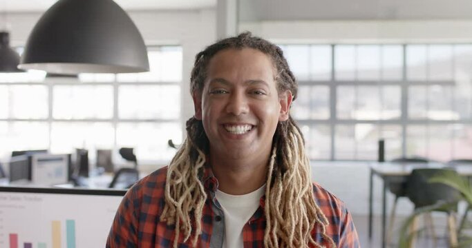 Portrait of happy biracial businessman with dreadlocks smiling in creative office in slow motion