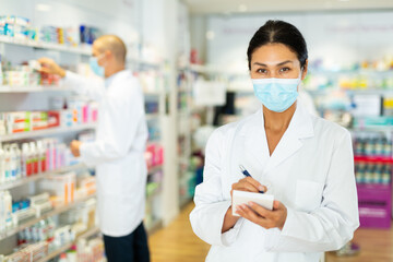 Asian female pharmacist in lab coat and face mask standing in salesroom of drugstore and writing recipe in blank.