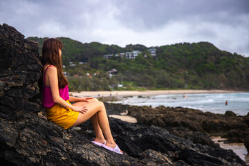 Beautiful girl stand on large rocks and admires powerful  waves in popular Byron Bay, NSW, Australia